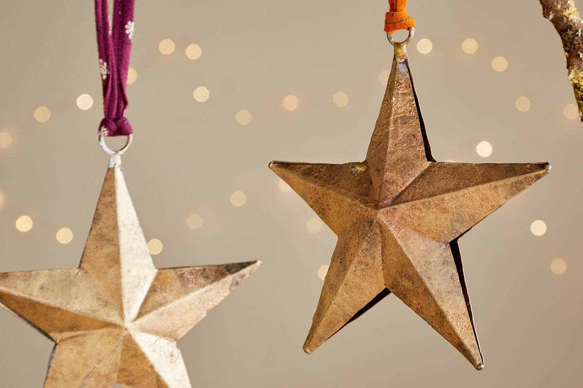 The nkuku Roshani Star Baubles - Antique Brass (Set of 2) feature metallic gold stars hanging from purple and orange ribbons, set against a soft beige backdrop with blurred lights for a festive, warm holiday ambiance.