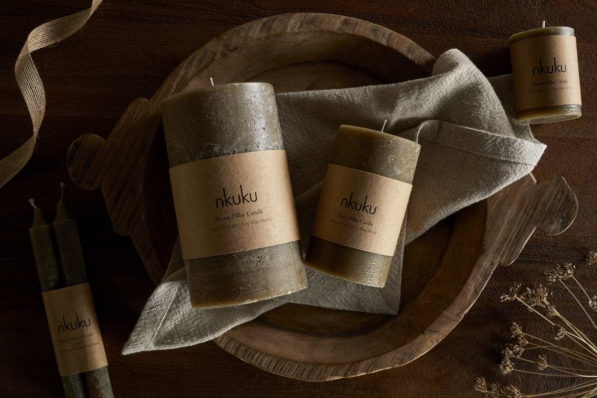 A rustic wooden tray holds three nkuku Rustic Soy Blend Mini Pillar Candles in Olive on a folded linen cloth, accented by two tapered candles and dried flowers atop a dark wooden surface.