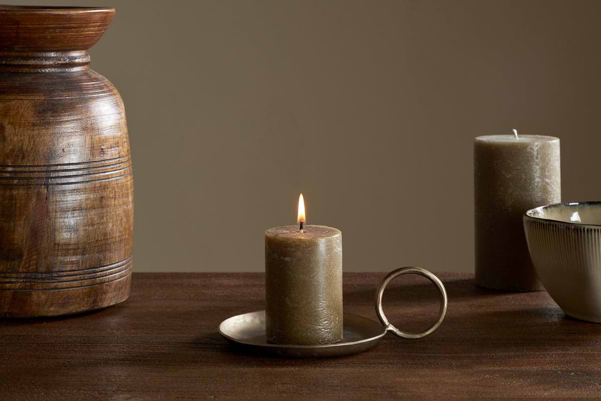 The nkuku Rustic Soy Blend Mini Pillar Candle in Olive glows on a brass handled holder atop a wooden surface, accompanied by a wooden vase, ceramic cup, and an unlit candle, all set against a beige background.