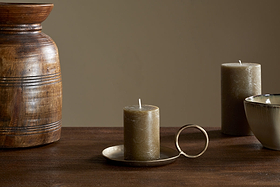 The nkuku Rustic Soy Blend Mini Pillar Candle in Olive sits on a brass holder with handle atop a wooden table, accompanied by a ceramic bowl, another candle, and a textured wooden vase against a plain beige background.