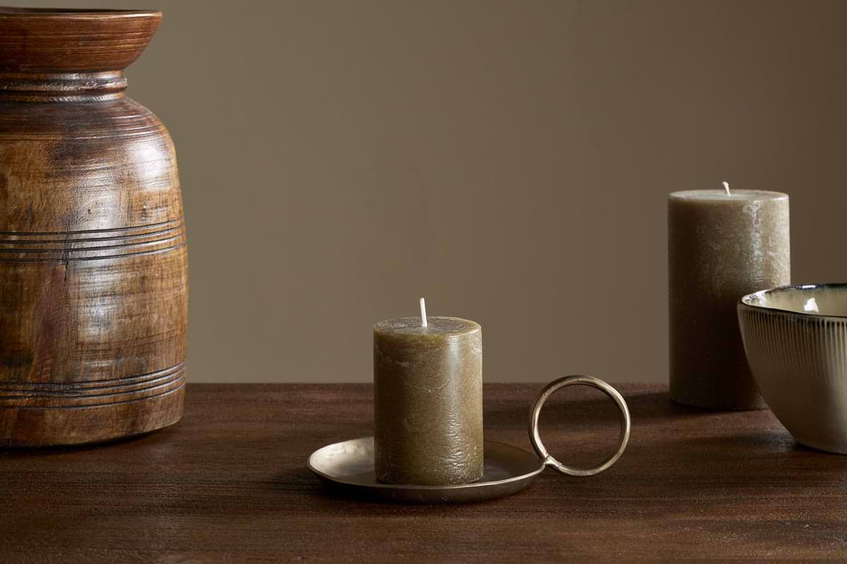 The nkuku Rustic Soy Blend Mini Pillar Candle in Olive sits on a brass holder with handle atop a wooden table, accompanied by a ceramic bowl, another candle, and a textured wooden vase against a plain beige background.