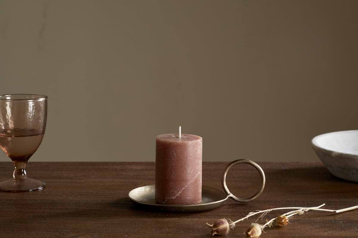 A nkuku Rustic Soy Blend Mini Pillar Candle in Rust with a textured finish sits on a brass handled holder atop a wooden table, beside a pink glass, white bowl, and dried flowers. The softly lit background is plain.