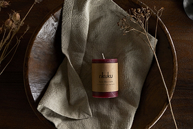 A Rustic Soy Blend Mini Pillar Candle - Dark Cherry by nkuku, with a textured finish, rests on a folded beige cloth in a large wooden bowl, surrounded by dried plants on a dark wooden surface.