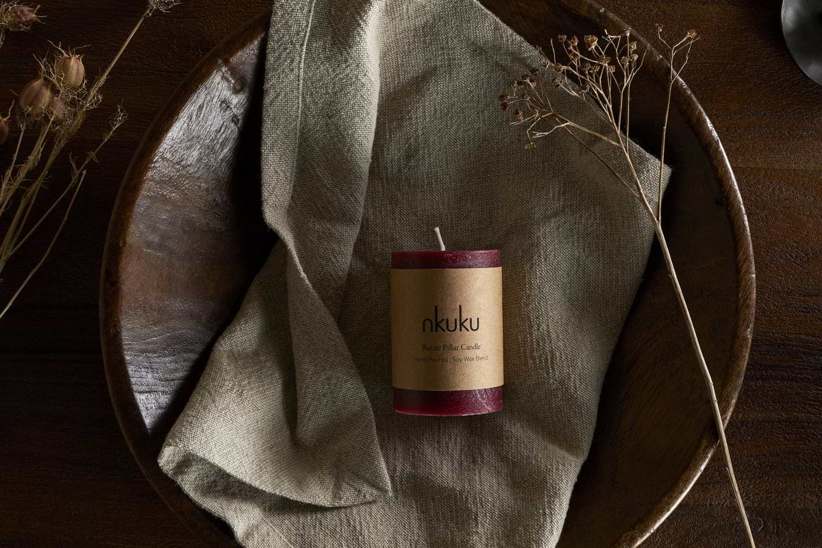 A Rustic Soy Blend Mini Pillar Candle - Dark Cherry by nkuku, with a textured finish, rests on a folded beige cloth in a large wooden bowl, surrounded by dried plants on a dark wooden surface.