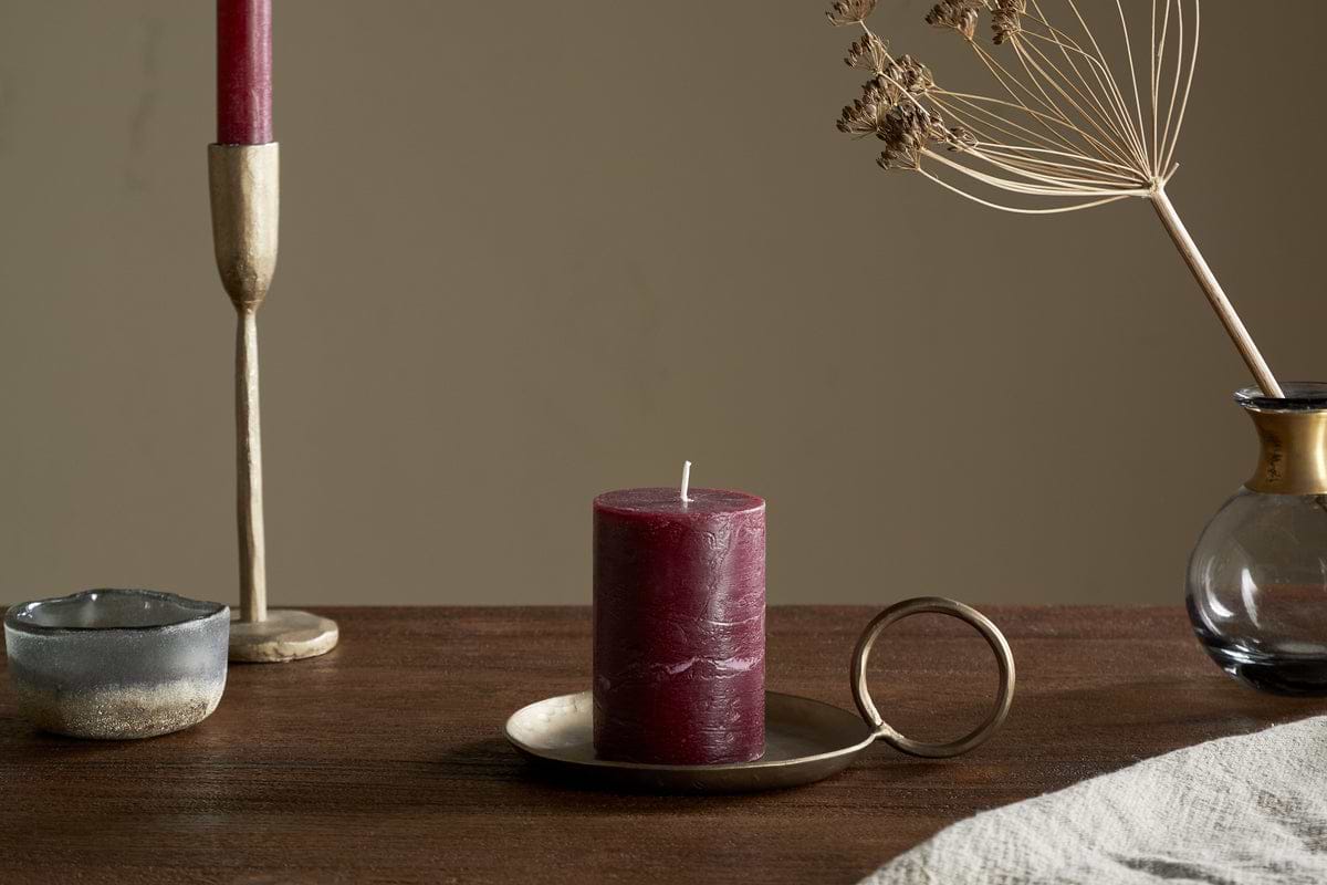 A Rustic Soy Blend Mini Pillar Candle in Dark Cherry by nkuku with a textured finish sits on a brass holder atop a wooden table, beside a gold taper holder with a red candle, a small ceramic bowl, and a glass vase of dried flowers.