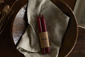 The nkuku Rustic Soy Blend Dinner Candles in Dark Cherry (Set of 2) feature a textured finish and come beautifully wrapped, displayed on a beige cloth in a rustic wooden bowl with dried flowers and an open book on a wooden surface.
