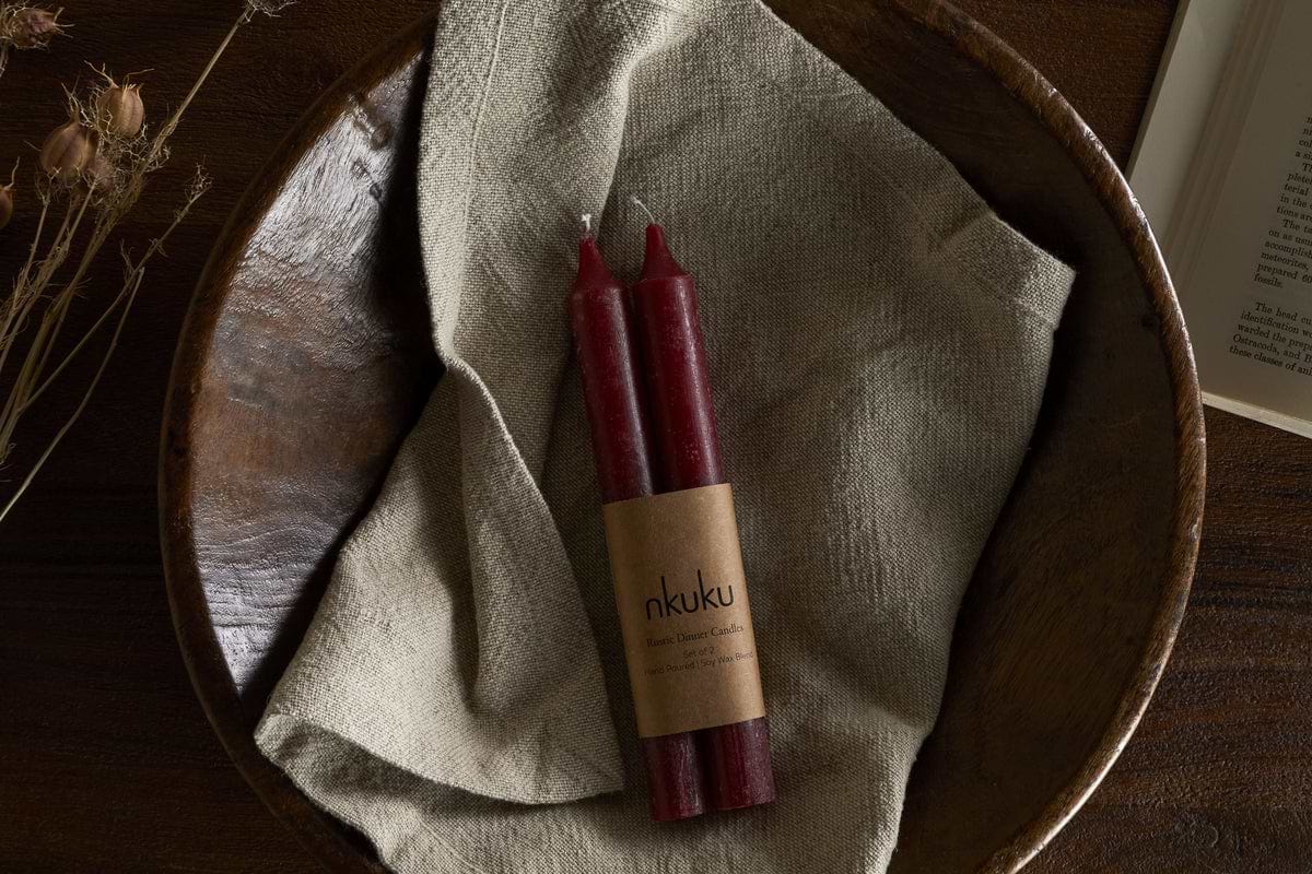 The nkuku Rustic Soy Blend Dinner Candles in Dark Cherry (Set of 2) feature a textured finish and come beautifully wrapped, displayed on a beige cloth in a rustic wooden bowl with dried flowers and an open book on a wooden surface.