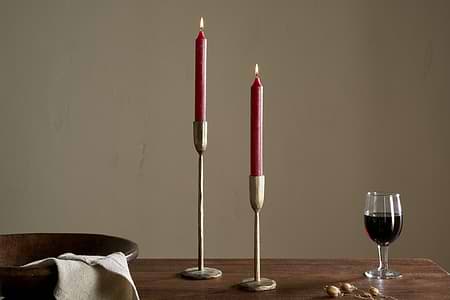 Two Rustic Soy Blend Dinner Candles in Dark Cherry by nkuku sit in brass holders on a wooden table, accompanied by a bowl, folded beige cloth, dried botanicals, and a glass of red wine against a plain beige wall.