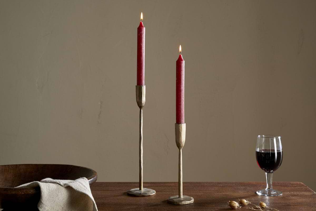 Two Rustic Soy Blend Dinner Candles in Dark Cherry by nkuku sit in brass holders on a wooden table, accompanied by a bowl, folded beige cloth, dried botanicals, and a glass of red wine against a plain beige wall.