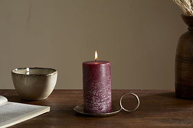 The nkuku Rustic Soy Blend Pillar Candle in Dark Cherry (Small) with a textured finish sits on a metal holder on a wooden table beside an open book, ceramic bowl, and brown vase with dried flowers in soft light.