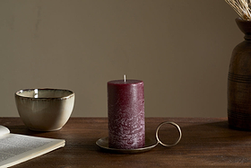 The nkuku Rustic Soy Blend Pillar Candle in Dark Cherry sits on a metal tray with a round handle on a wooden table alongside an open book, ceramic bowl, and brown vase with dried grasses.