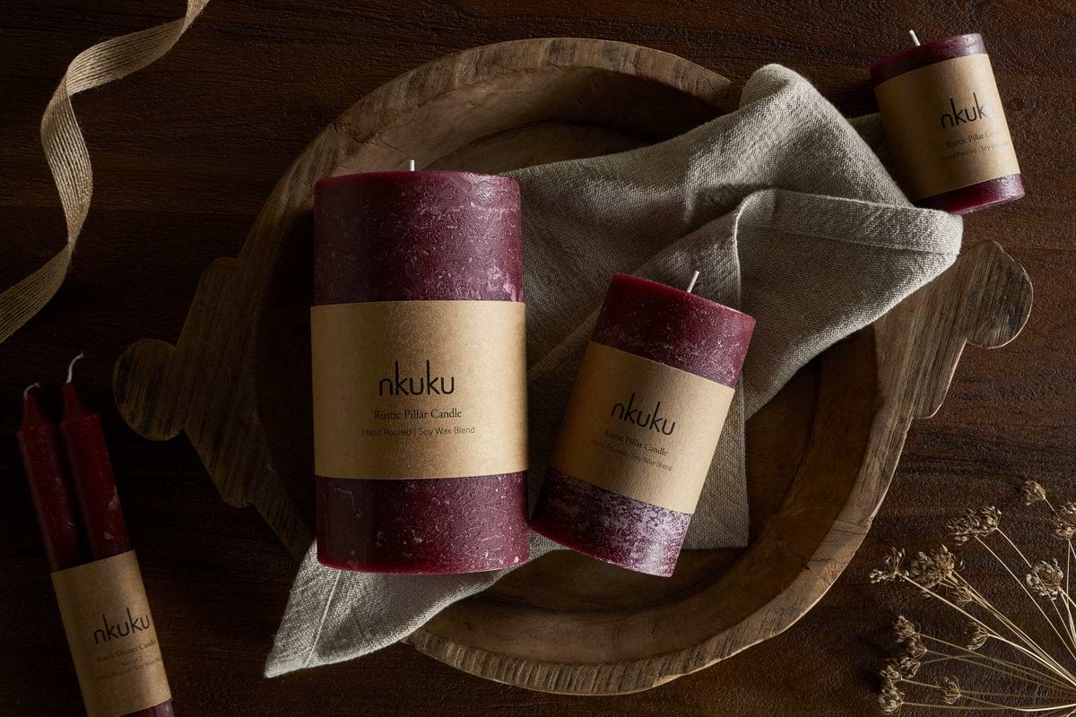 A wooden bowl with a beige napkin holds two nkuku Rustic Soy Blend Pillar Candles in Dark Cherry. Nearby, a small and a tall textured nkuku candle rest on wood amid dried flowers and ribbon.