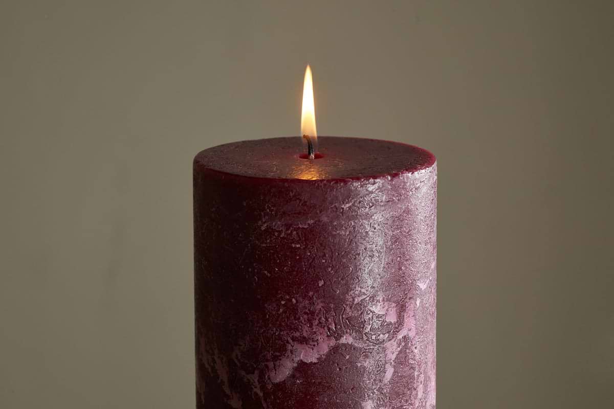 The nkuku Rustic Soy Blend Pillar Candle - Dark Cherry - Large glows softly with a steady flame, its textured finish in rich cherry hues standing out against a simple, light background.