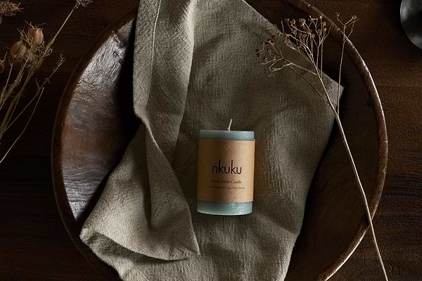 A Rustic Soy Blend Mini Pillar Candle in Sage Green by nkuku, featuring a textured finish, rests on a beige cloth in a wooden bowl with dried plants, atop a dark wooden surface.