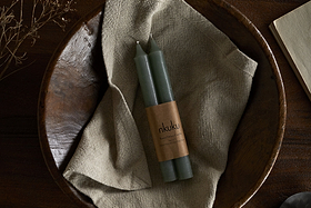 A set of two nkuku Rustic Soy Blend Dinner Candles in sage green, wrapped in a branded label, rest on beige cloth in a wooden bowl, with a dried plant and part of an open book on a dark wooden surface.