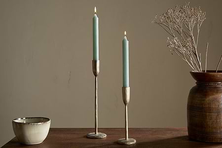 A wooden table features nkukus Rustic Soy Blend Dinner Candle - Sage Green (Set of 2) in brass holders, a ceramic bowl, and a brown vase with dried branches, all set against a beige wall.