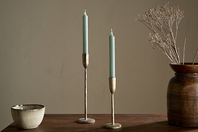 A wooden table with a cream bowl, a brown vase with dried branches, and two nkuku Rustic Soy Blend Dinner Candles in sage green held by brass candlesticks, set against a plain beige wall.
