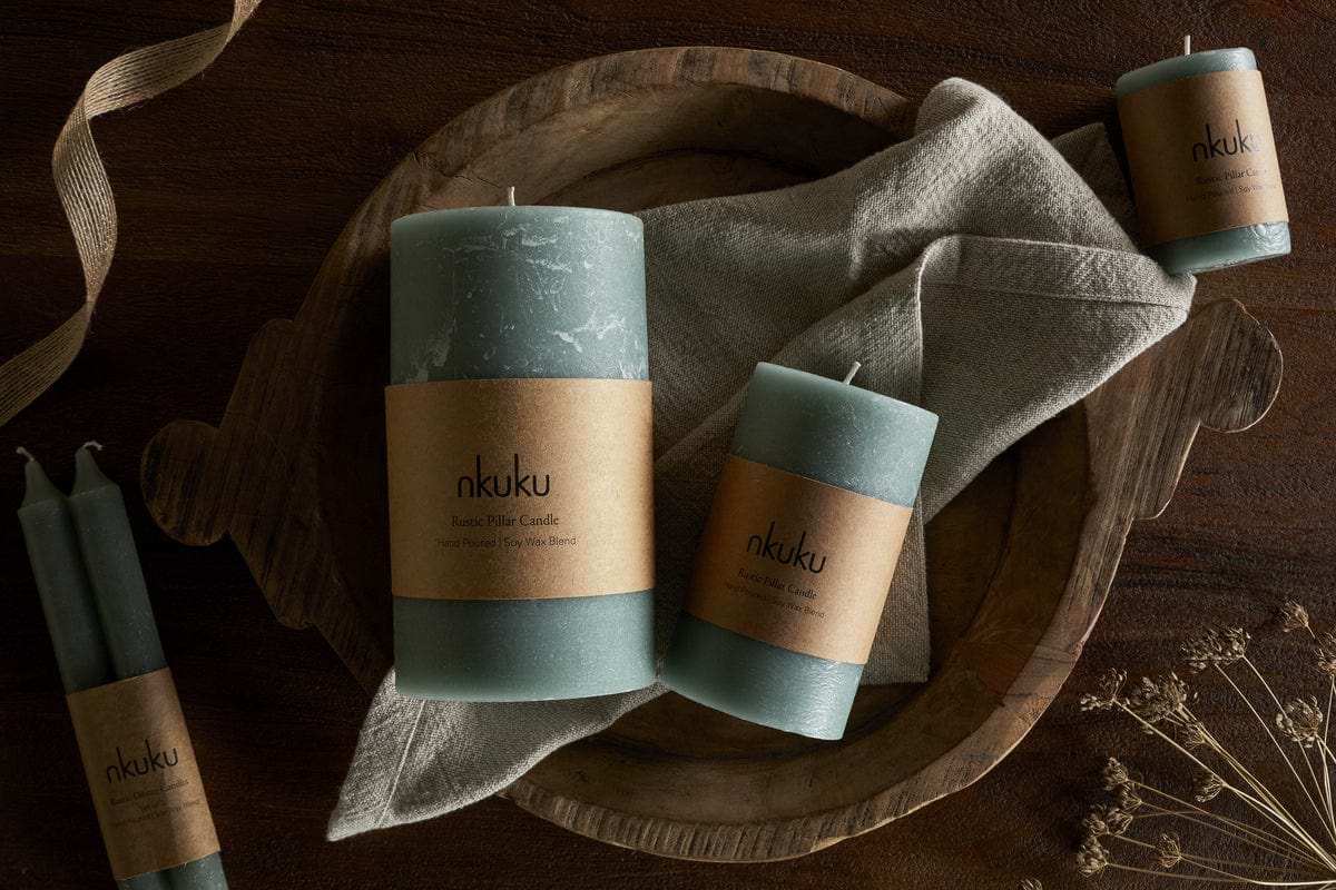 A wooden tray holds two nkuku Rustic Soy Blend Pillar Candles in Sage Green (Small) with brown paper labels, set on a beige cloth. Two smaller candles and dried flowers rest nearby on the dark wooden surface.