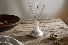 The nkuku Cardamom & Amber Reed Diffuser with wooden reeds rests on a rustic table, its scent of sandalwood and cardamom blending with dried fruits and a folded beige cloth for an inviting display.