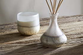 A rustic wooden table features the nkuku Cardamom & Amber Reed Diffuser, its natural sticks releasing warm, soothing notes for a calm, minimalistic atmosphere.