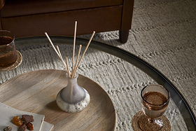 A glass coffee table with a wooden tray displays the nkuku Cardamom & Amber Reed Diffuser, a cloth napkin with small stones, and two amber glasses on woven coasters. Beneath it is a patterned beige rug.