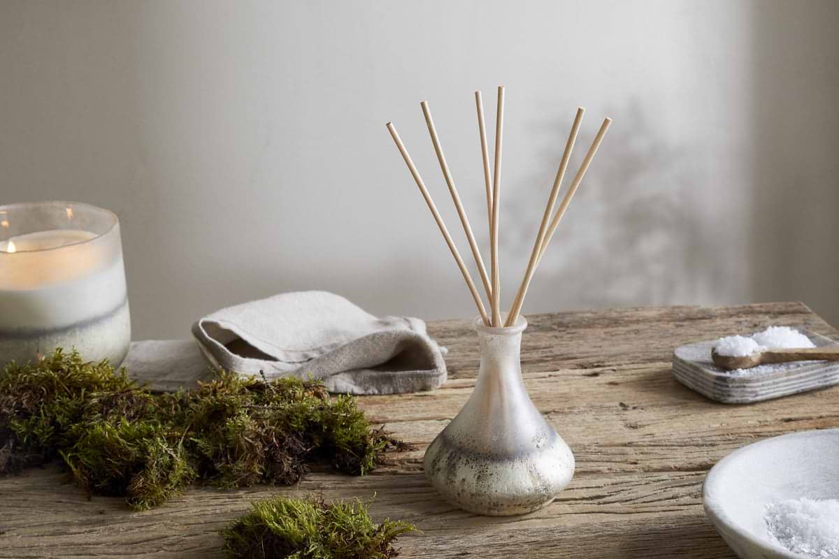 A nkuku Sea Salt & Moss Reed Diffuser sits in a small glass vase on a rustic wooden table, surrounded by green moss, a lit candle, a folded cloth, and a bowl of salt with a wooden spoon, creating an atmosphere of calm and natural beauty.