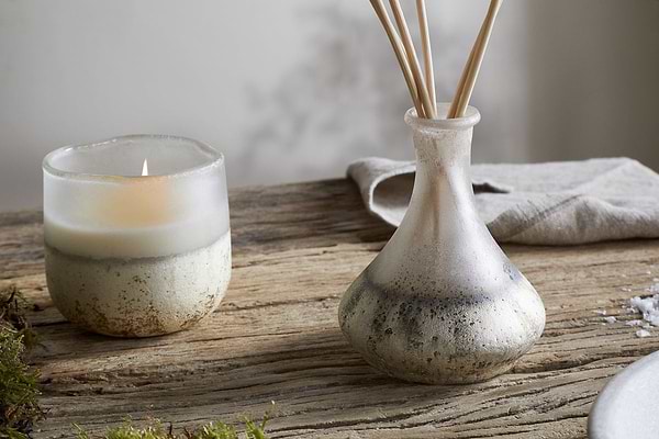 A lit candle in a frosted glass holder and the nkuku Sea Salt & Moss Reed Diffuser with slender reeds sit on rustic wood, alongside moss and a folded cloth, creating an inviting scene enhanced by a bespoke fragrance.