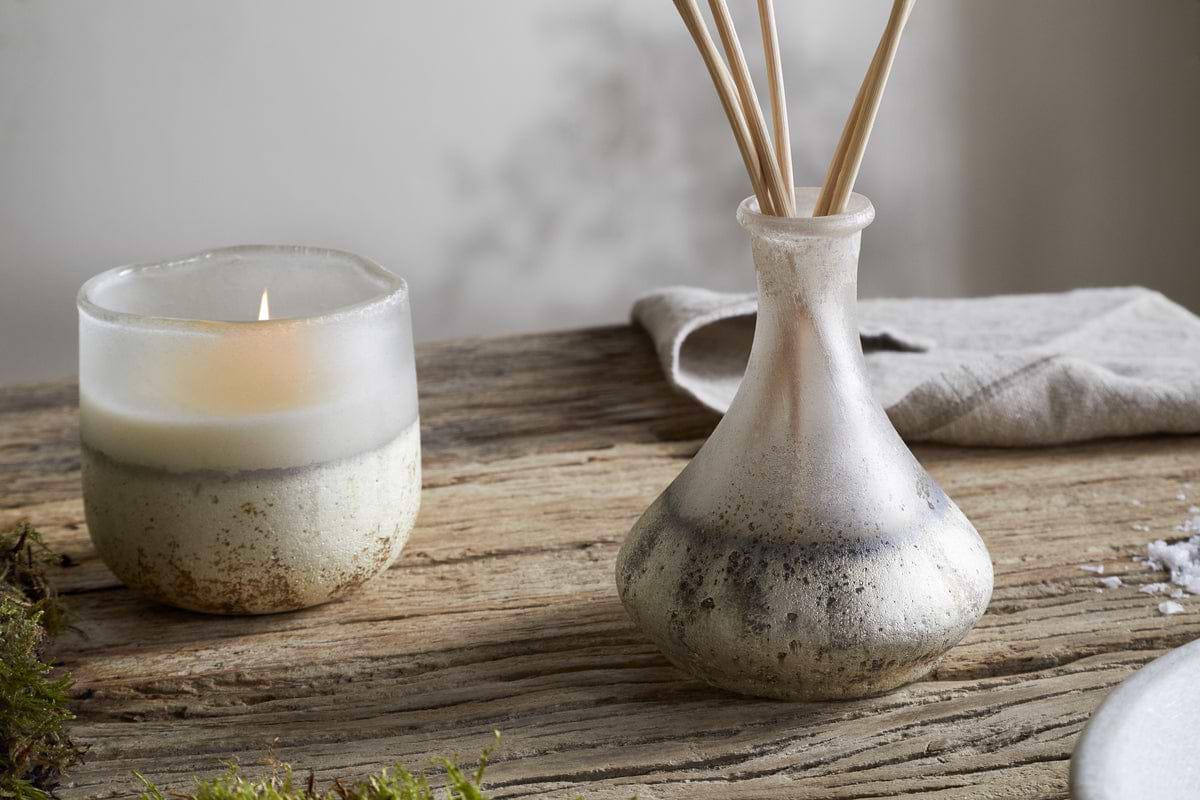 A lit candle in a frosted glass holder and the nkuku Sea Salt & Moss Reed Diffuser with slender reeds sit on rustic wood, alongside moss and a folded cloth, creating an inviting scene enhanced by a bespoke fragrance.