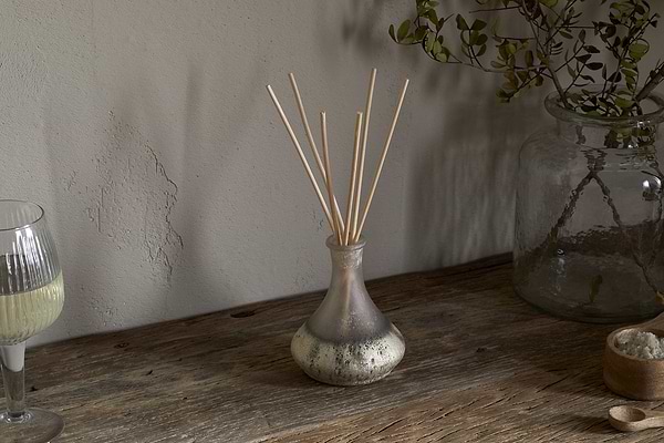 The nkuku Sea Salt & Moss Reed Diffuser adds a bespoke scent to a rustic wooden table, alongside a partial glass of white wine, a large jar with foliage, and a small bowl of salts.