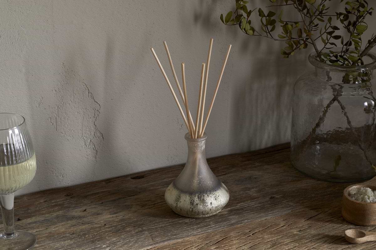 The nkuku Sea Salt & Moss Reed Diffuser adds a bespoke scent to a rustic wooden table, alongside a partial glass of white wine, a large jar with foliage, and a small bowl of salts.