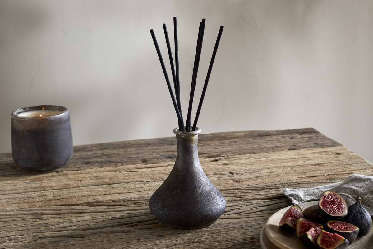 A nkuku Cedarwood & Fig Reed Diffuser with black reeds stands on a rustic wooden table, accompanied by a dark candle and fresh figs, infusing the air with a cozy natural cedarwood scent.