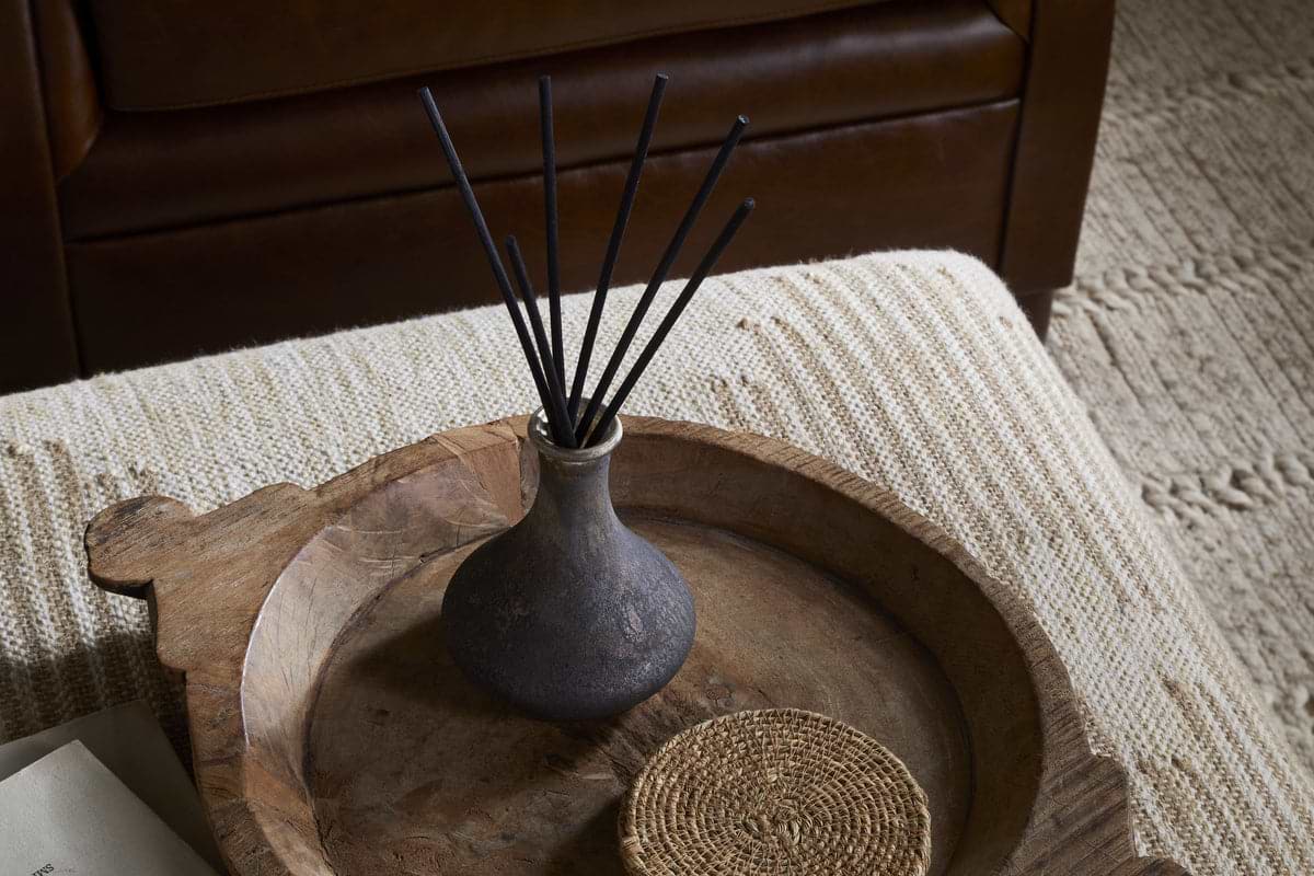 The nkuku Cedarwood & Fig Reed Diffuser, with black reeds in a dark ceramic vase, sits on a rustic wooden tray beside a woven coaster atop a textured ottoman, filling the air with cedarwood aroma.