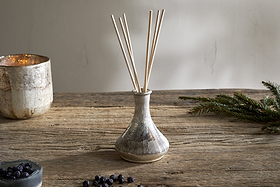 The nkuku Juniper & Fir Reed Diffuser, with reed sticks in a ceramic vase, sits on a rustic wooden table alongside an evergreen branch, scattered berries, a metallic candle holder, and a small bowl for a stylish setting.