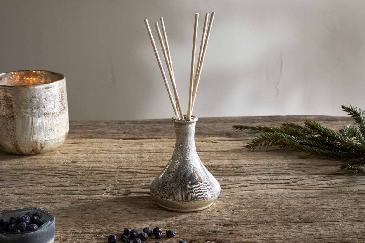 The nkuku Juniper & Fir Reed Diffuser, with reed sticks in a ceramic vase, sits on a rustic wooden table alongside an evergreen branch, scattered berries, a metallic candle holder, and a small bowl for a stylish setting.