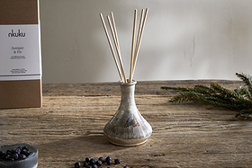 A rustic nkuku Juniper & Fir Reed Diffuser with bespoke fragrance sits on a wooden table, surrounded by sprigs of fir and juniper berries. In the background, a box reads “nkuku Juniper & Fir.”.