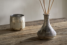 A rustic wooden table displays a metallic cup and a textured silver nkuku Juniper & Fir Reed Diffuser, its fragrance reeds set against a plain, light-colored background.