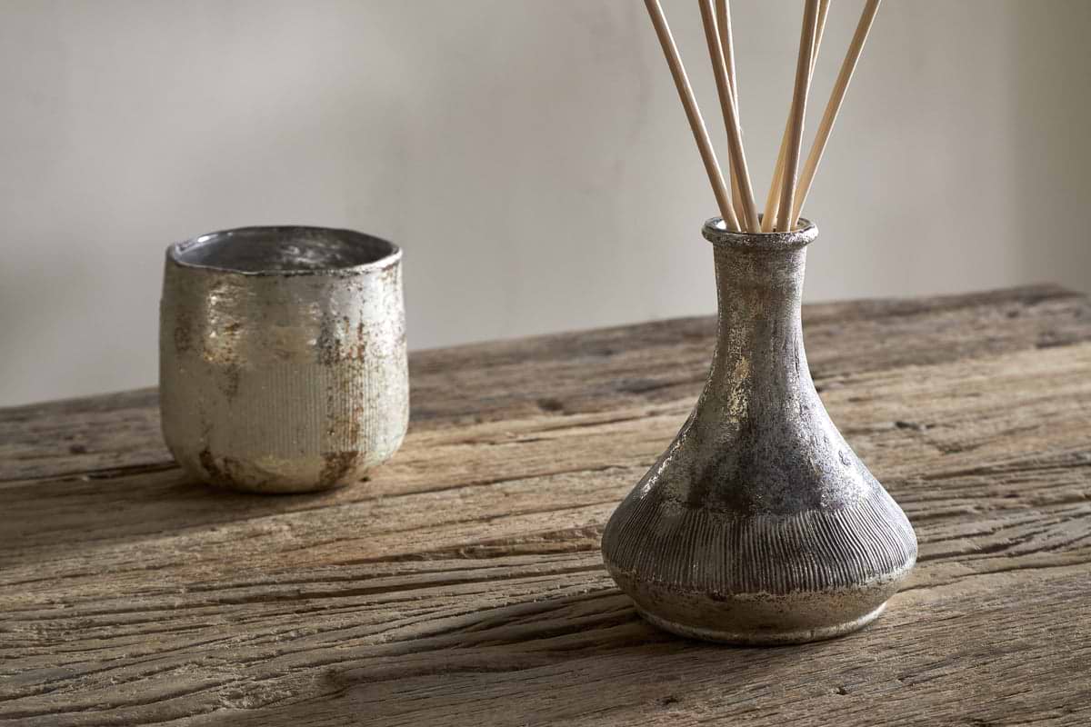 A rustic wooden table displays a metallic cup and a textured silver nkuku Juniper & Fir Reed Diffuser, its fragrance reeds set against a plain, light-colored background.