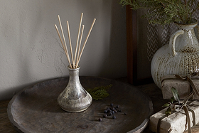 A nkuku Juniper & Fir Reed Diffuser in a rustic ceramic vase sits on a large wooden tray with wrapped gifts, brown ribbon, and greenery. A white ceramic vase with foliage is in the background on a wooden surface.