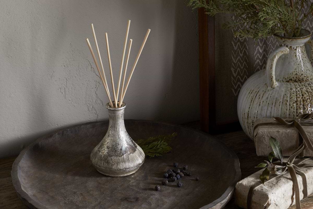 A nkuku Juniper & Fir Reed Diffuser in a rustic ceramic vase sits on a large wooden tray with wrapped gifts, brown ribbon, and greenery. A white ceramic vase with foliage is in the background on a wooden surface.