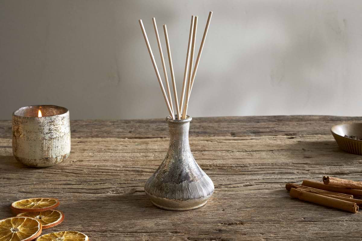 The nkuku Mandarin & Clove Reed Diffuser, with several reeds, sits on a rustic wooden table beside a textured candle holder, dried orange slices, and cinnamon sticks, filling the space with a cozy spiced fragrance.