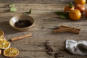 The nkuku Mandarin & Clove Reed Diffuser is pictured on a rustic wooden surface with dried orange slices, cinnamon sticks, fresh oranges, and cloves, evoking a festive and spiced mandarin fragrance.