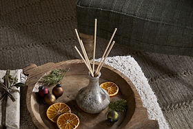A wooden tray holds nkuku’s Mandarin & Clove Reed Diffuser, filling the air with festive scent, while dried orange slices, greenery, and ornaments lie on a textured rug beside a green upholstered chair.