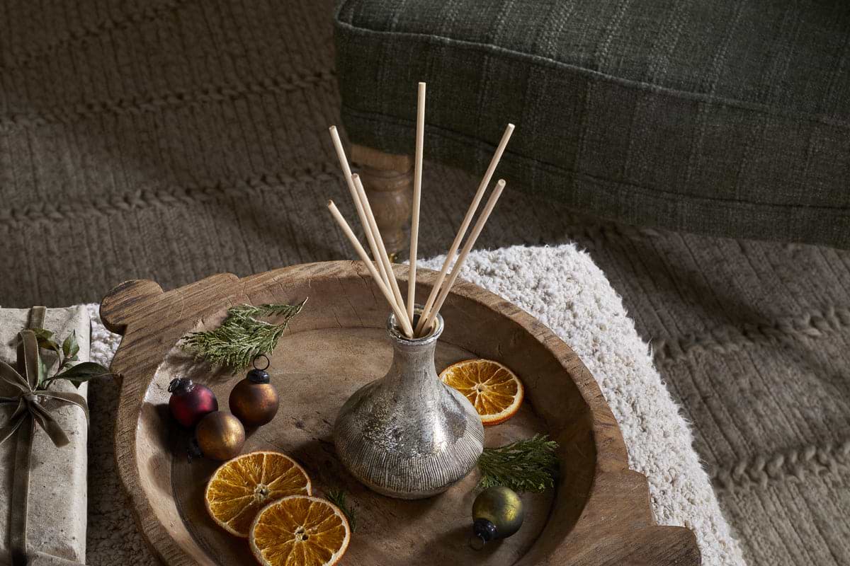 A wooden tray holds nkuku’s Mandarin & Clove Reed Diffuser, filling the air with festive scent, while dried orange slices, greenery, and ornaments lie on a textured rug beside a green upholstered chair.