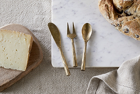 A block of cheese, bread, a beige napkin, and the nkuku Roda Serving Set – Antique Brass (Set of 3) on marble and linen create an elegant display.