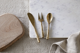 The nkuku Roda Serving Set - Antique Brass (set of 3) is displayed on a white marble slab next to a wooden cutting board and a textured beige cloth, all set atop a fabric surface.
