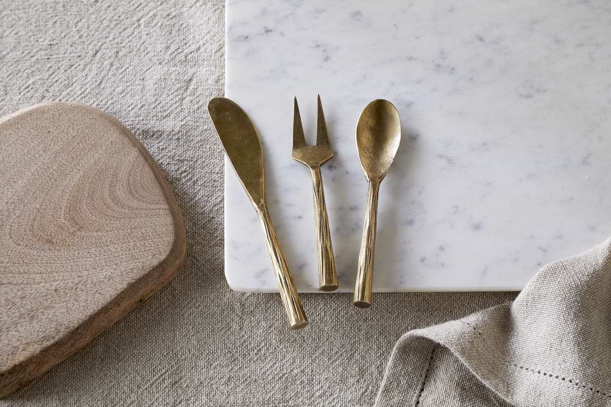 The nkuku Roda Serving Set - Antique Brass (set of 3) is displayed on a white marble slab next to a wooden cutting board and a textured beige cloth, all set atop a fabric surface.
