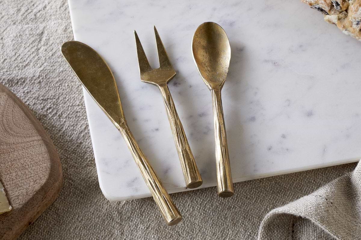 The nkuku Roda Serving Set – Antique Brass (Set of 3), featuring a spreader, fork, and spoon, rests on white marble beside bread atop beige fabric.