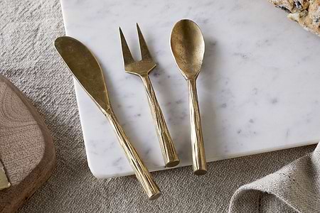 The nkuku Roda Serving Set – Antique Brass (Set of 3), featuring a spreader, fork, and spoon, rests on white marble beside bread atop beige fabric.