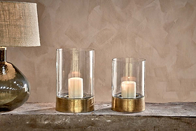 Two Rajura Hurricane Lanterns by nkuku with gold bases rest on a rustic wooden surface beside a glass-based lamp with a textured beige shade, all set against a textured beige wall, creating warm and cozy ambient lighting.