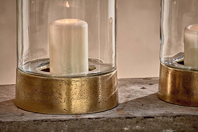 The Rajura Hurricane Lantern by nkuku features a white pillar candle in a cylindrical glass with a textured brass-toned base, set on rustic wood. Its warm glow creates an inviting atmosphere, with another Rajura lantern partially visible to the right.